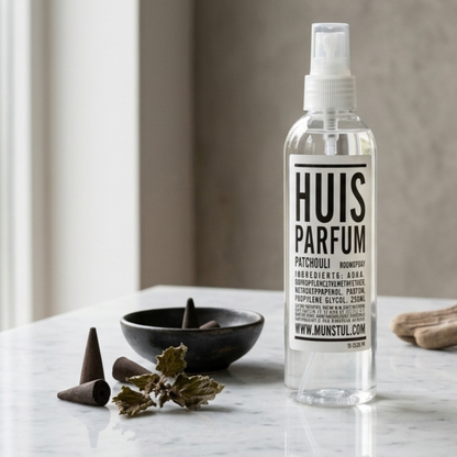 Mijn Stijl roomspray Patchouli (250 ml) in een transparante fles met verstuiver, staand op een licht, marmeren oppervlak. Links staat een klein, donker schaaltje met wierookkegeltjes, met daarvoor nog een paar losse kegeltjes en gedroogde blaadjes. Rechtsachter zijn vage contouren van houten accessoires zichtbaar tegen een achtergrond van sfeervolle, beige gordijnen.