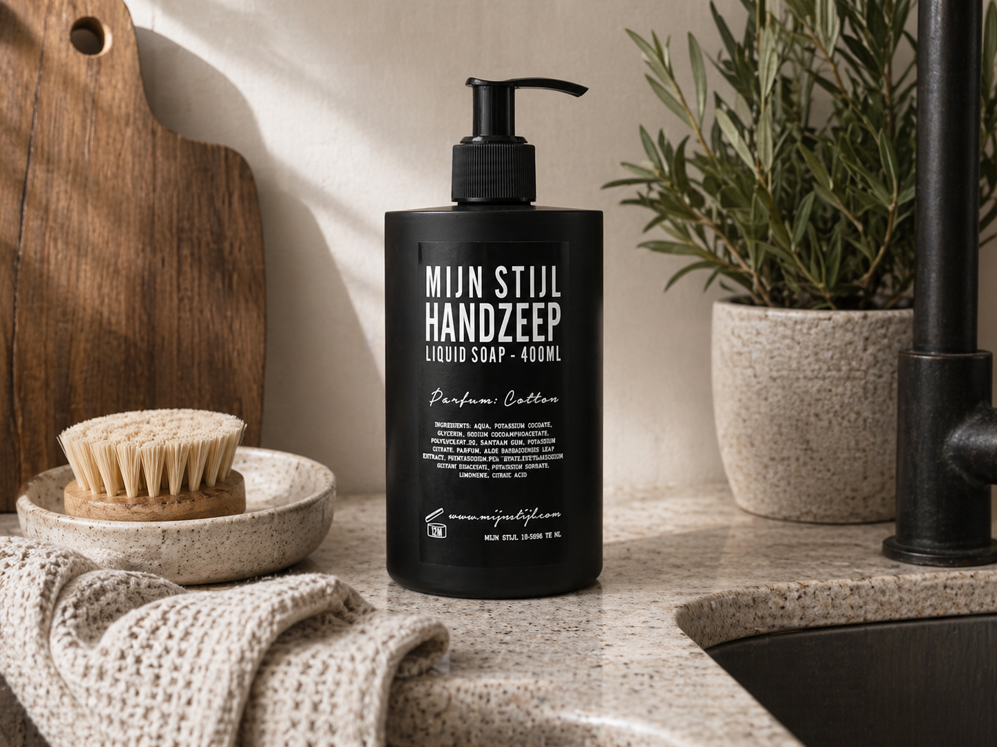 Mijn Stijl handzeep Cotton (400 ml) in een zwarte fles met pompje, geplaatst op een licht natuurstenen aanrechtblad naast een donkere kraan en spoelbak. Links op de voorgrond ligt een beige handdoek met wafelstructuur, met daarachter een klein rond schaaltje met een houten afwasborstel en een houten snijplank tegen de muur. Rechtsachter staat een lichte, gespikkelde pot met een groene plant.