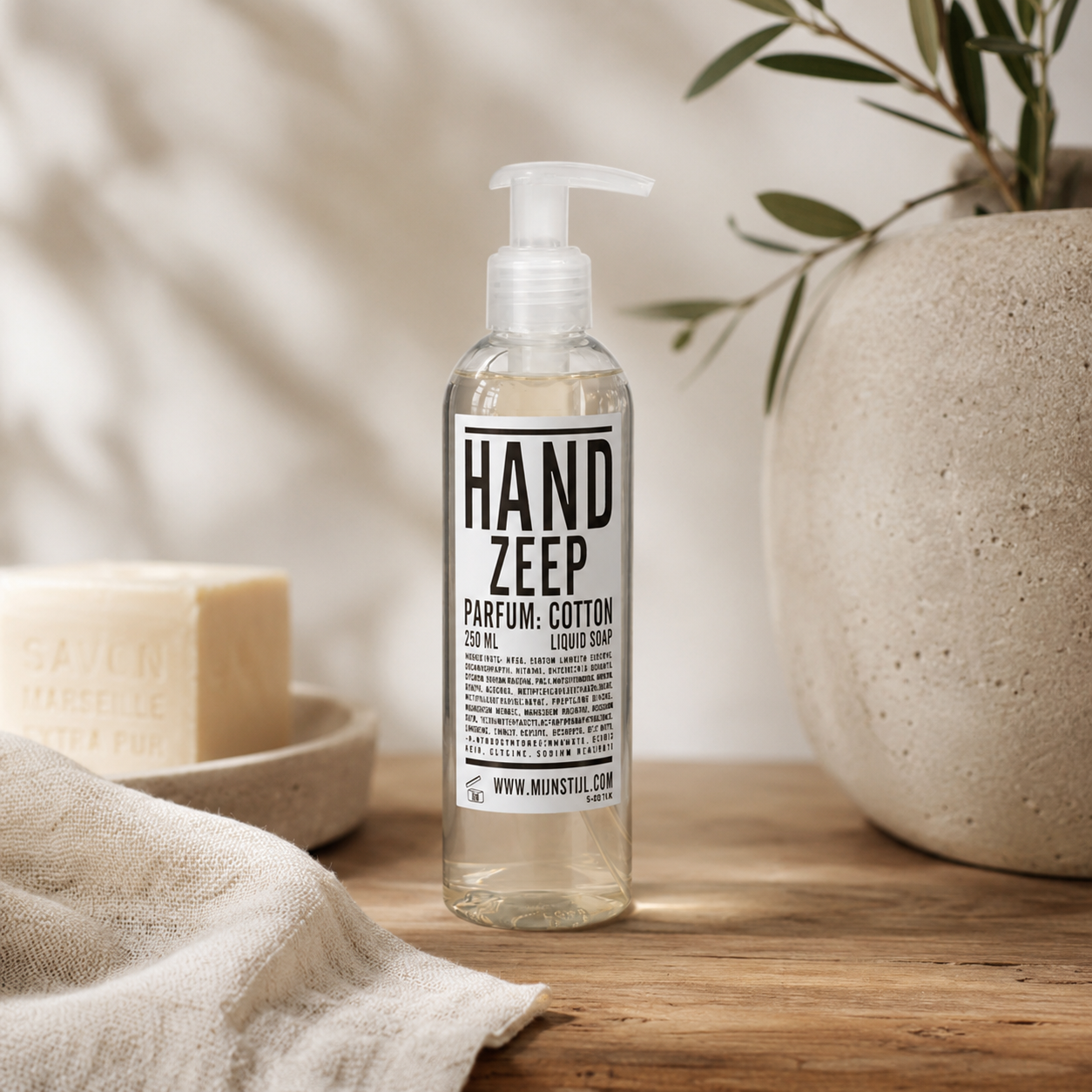 Mijn Stijl handzeep Cotton (250 ml) in een transparante fles, centraal geplaatst op een robuuste houten plank. Links op de voorgrond ligt een beige, geweven handdoek en daarachter staat een klein, rond schaaltje met een blokzeep. Rechts staat een grote, beige aardewerken pot of vaas met subtiele groene takken, tegen een lichte wand.