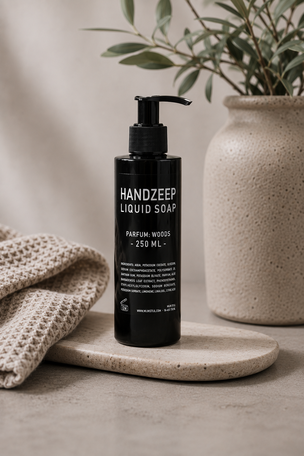 Mijn Stijl handzeep Woods in een zwarte fles met pompje in een natuurlijke setting, met een grofgebreide linnen doek links, een houten massageborstel rechts en een vaas met olijftakken op de achtergrond.