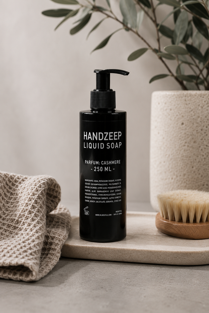 Mijn Stijl handzeep Cashmere in een zwarte fles met pompje in een natuurlijke setting, met een grofgebreide linnen doek links, een houten massageborstel rechts en een vaas met olijftakken op de achtergrond.